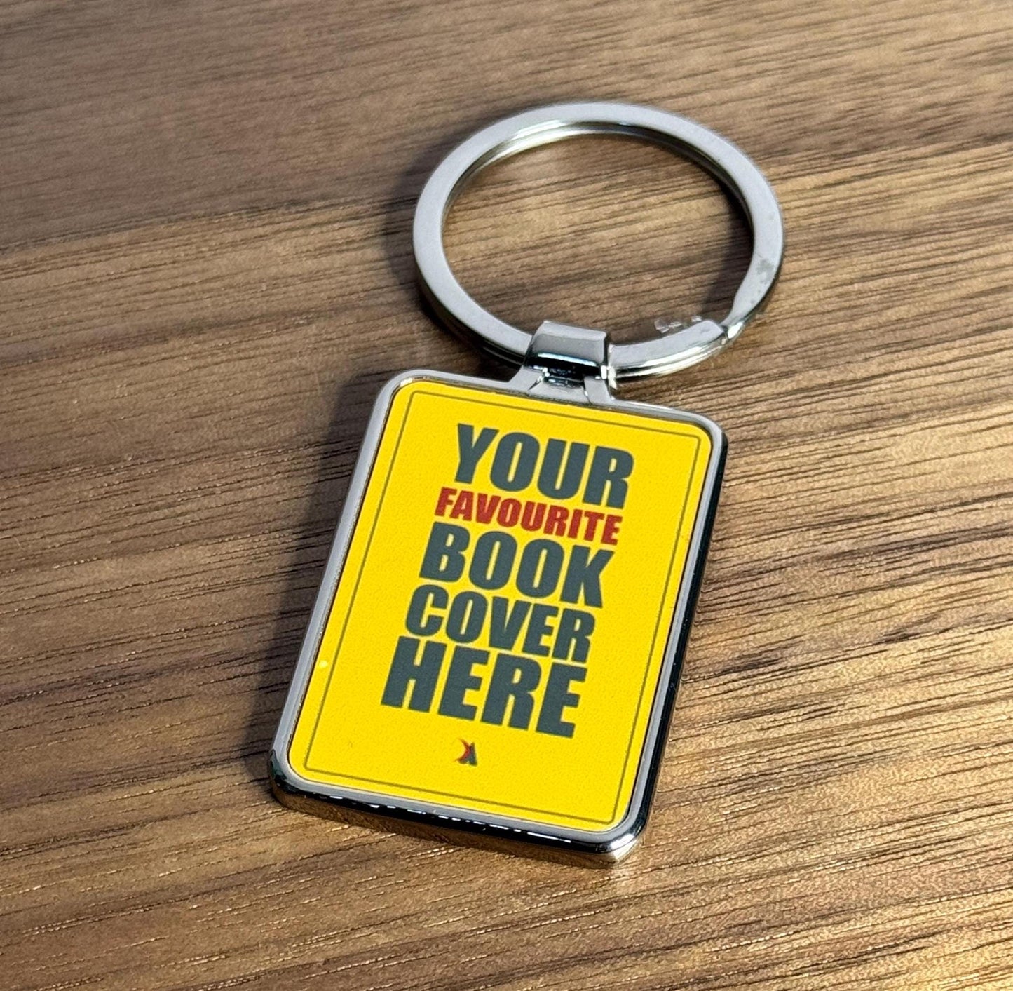 A silver keyring with an example book cover design. The design is yellow with the phrase ‘Your favourite book cover here’ in black.