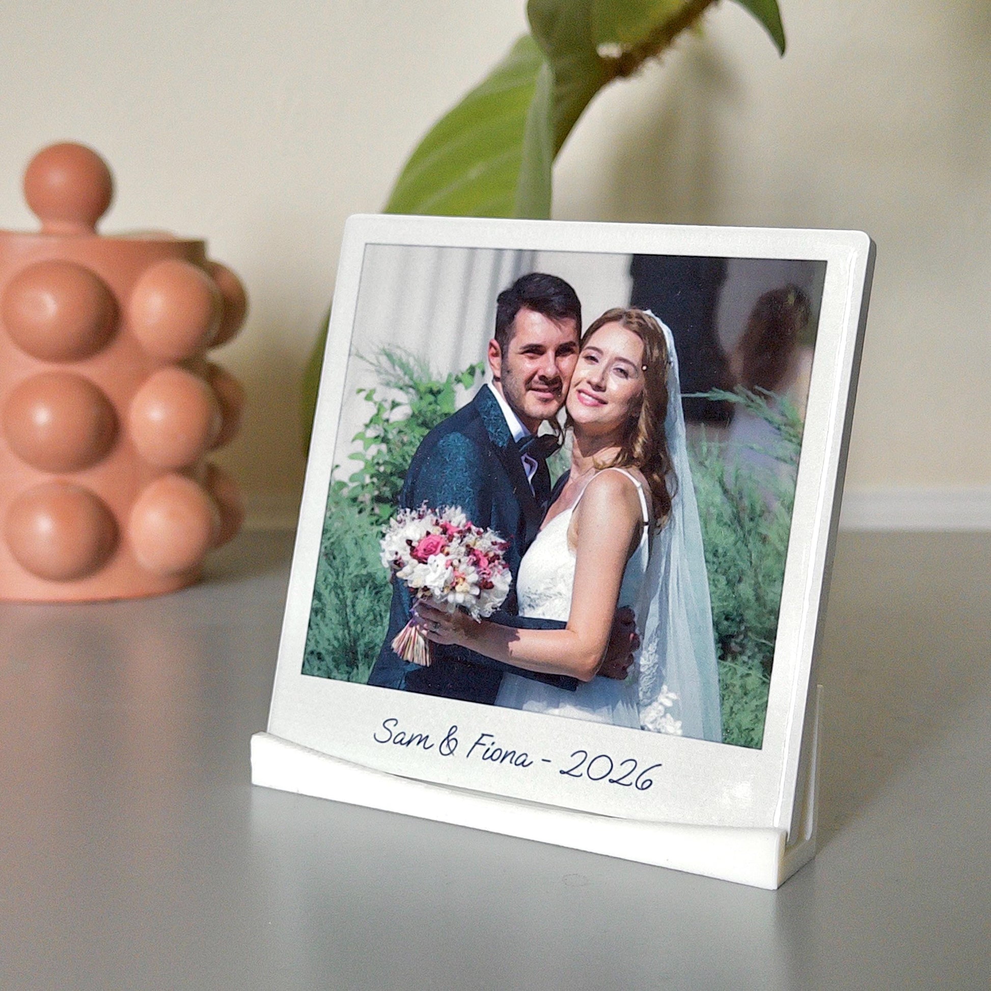 A ceramic tile printed with a wedding photo. There is a caption that says ’Sam and Fiona - 2026’. The tile is upright in a display stand.